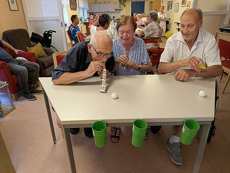 Seniorinnen und Senioren sitzen an Tisch und bewegen weißen Tennisball durch pusten in grüne Plastikbecher