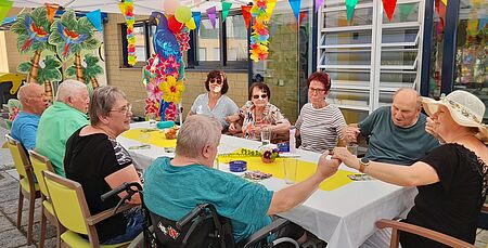 Seniorinnen, Senioren und Pflegekräfte sitzen um einen Tisch herum. Der Pavillon, unter dem sie sitzen, ist sommerlich geschmückt.