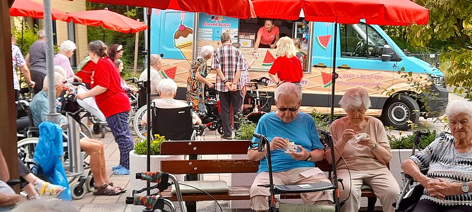 Eismobil "Meloni" Im Hintergrund blaues Eismobil, im Vordergrund Seniorinnen und Senioren, die Eis essen