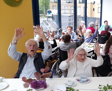 Senioren an Tischen heben ausgelassen die Hände zur Musik und singen mit