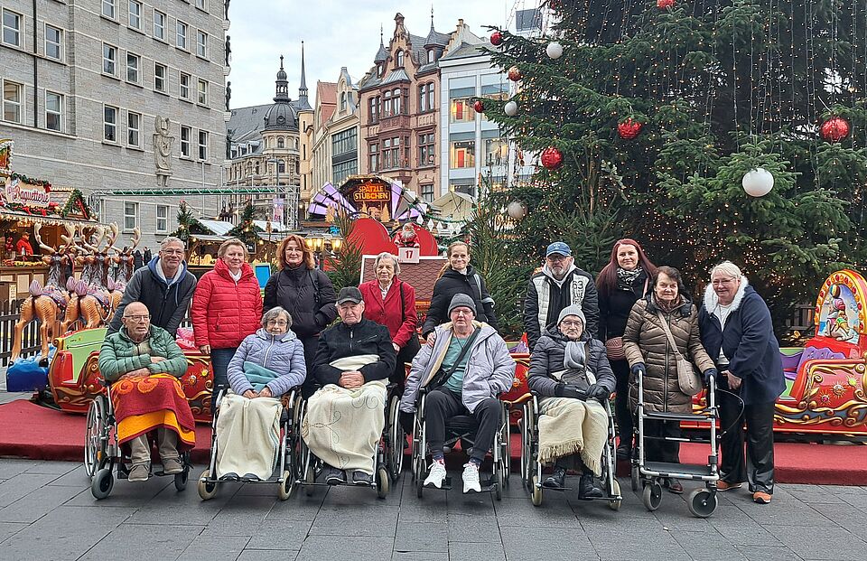 Seniorinnen und Senioren mit Betreuern und Angehörigen vor großem Tannenbaum und verschiednenen Hütten auf dem Weihnachtsmarkt