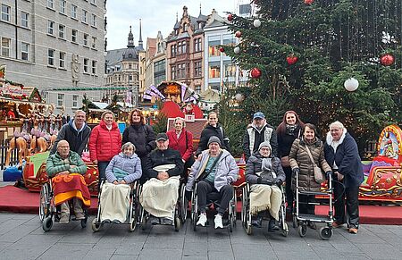 Seniorinnen und Senioren mit Betreuern und Angehörigen vor großem Tannenbaum und verschiednenen Hütten auf dem Weihnachtsmarkt