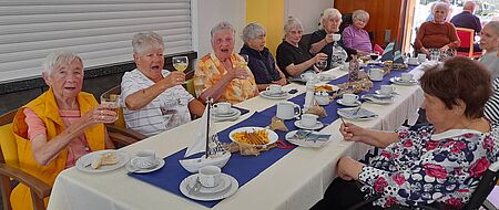Seniorinnen und Senioren sitzen um gedeckten Tisch herum und stoßen mit Gläsern an