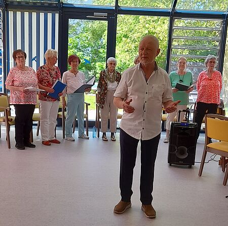 Mann in Hemd steht im Vordergrund und dirigiert, im Hintergrund sind Seniorinnen zu sehen, die singen