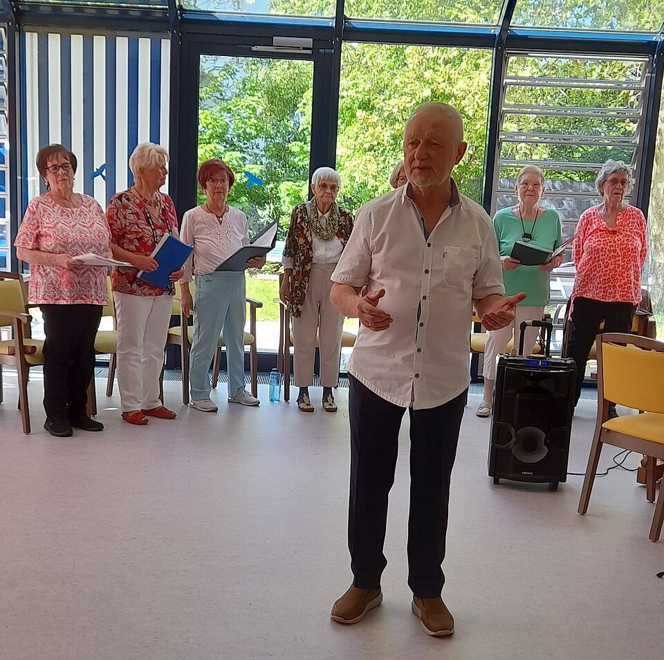 Mann in Hemd steht im Vordergrund und dirigiert, im Hintergrund sind Seniorinnen zu sehen, die singen
