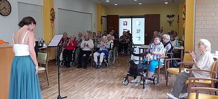 Sängerin in weißem Oberteil und grünem Rock steht vor Publikum, bestehend aus Seniorinnen und Senioren, die begeistert klatschen.