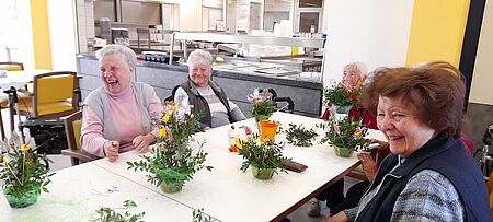 Seniorinnen sitzen um Tisch verteilt, vor ihnen grüne Gestecke, und lachen