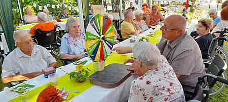 Seniorinnen und Senioren sitzen in geschmücktem Garten an Tisch und schauen auf Glücksrad, das vor ihnen steht