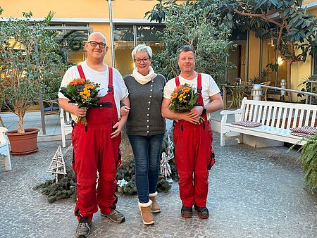 Hausmeister in roten Overalls mit Blumensträußen, in der Mitte Einrichtungsleiterin, alle schauen in Kamera