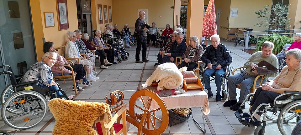 Seniorinnen und Senioren sitzen im Kreis um Spinnrad herum
