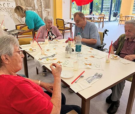 Seniorinnen und Senioren sitzen um Tisch herum verteilt und probieren kleine Snacks