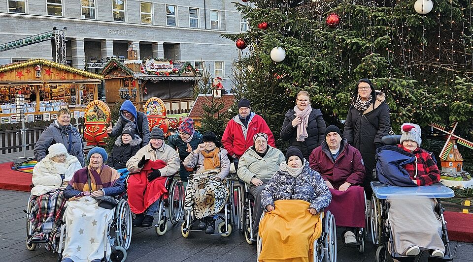 Seniorinnen und Senioren mit Betreuern vor großem Weihnachtsbaum, im Hintergrund verschiedene Verkaufsstände