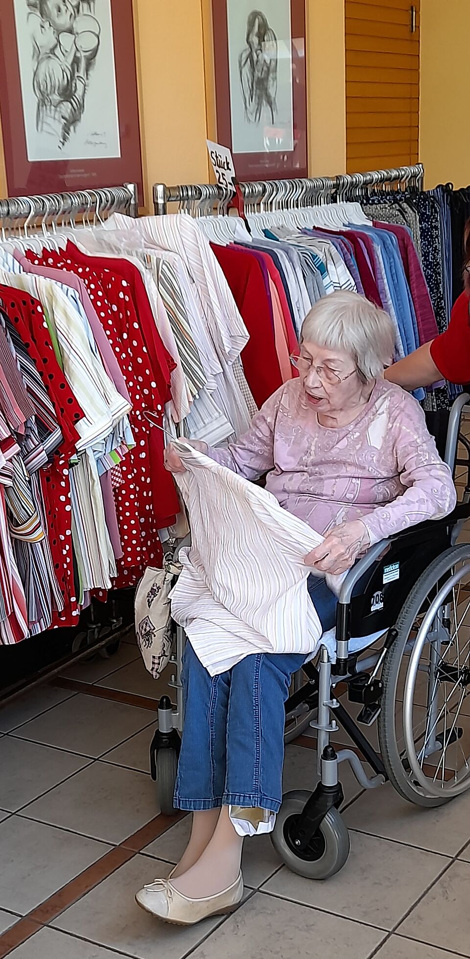 Seniorin in Rollstuhl stöbert durch Kleidung an Kleiderständer