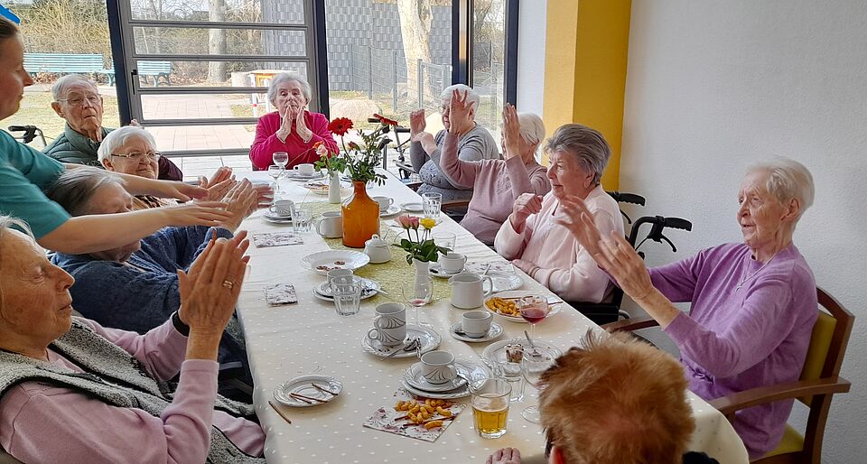 Seniorinnen sitzen an langer, gedeckter Tafel und stoßen mit Gläsern an