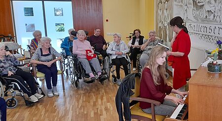 Frau an Klavier im Vordergrund, daneben Frau, die aus Buch vorliest. Im Hintergrund sitzen Seniorinnen und Senioren